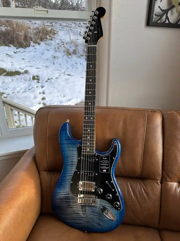 A blue Fender American Ultra Stratocaster electric guitar with a flame maple top is propped upright on a brown leather…