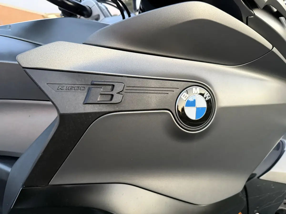 A close-up photo of a matte gray BMW motorcycle's fuel tank, showing the embossed 'K1600 B' model name and the iconic…