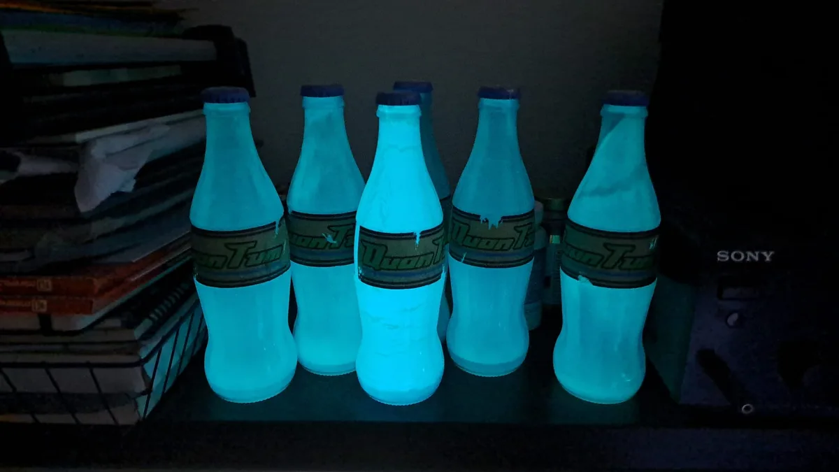 In a dark room, six glass bottles with glowing blue liquid and labels reading 'QuonTum' are lined up on a shelf. To the left…