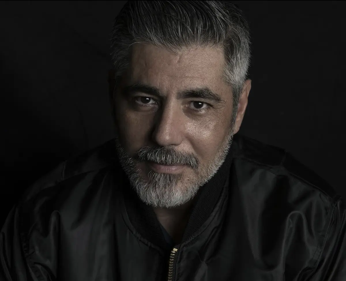a close-up portrait of george clooney with gray hair and beard, wearing a black bomber jacket against a dark background. The…