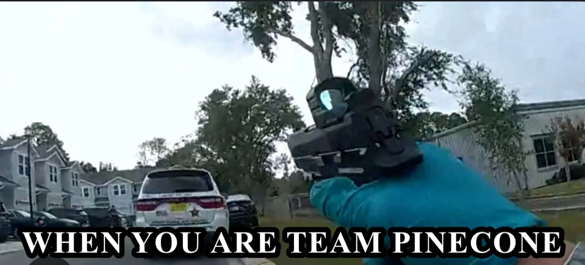A first-person view shows a hand holding a handgun with a red dot sight, aimed forward in a residential street. A white…
