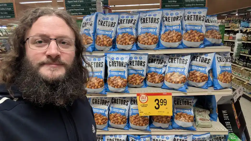 A man with long curly hair, glasses, and a full beard stands in a grocery store aisle in front of shelves stocked with…