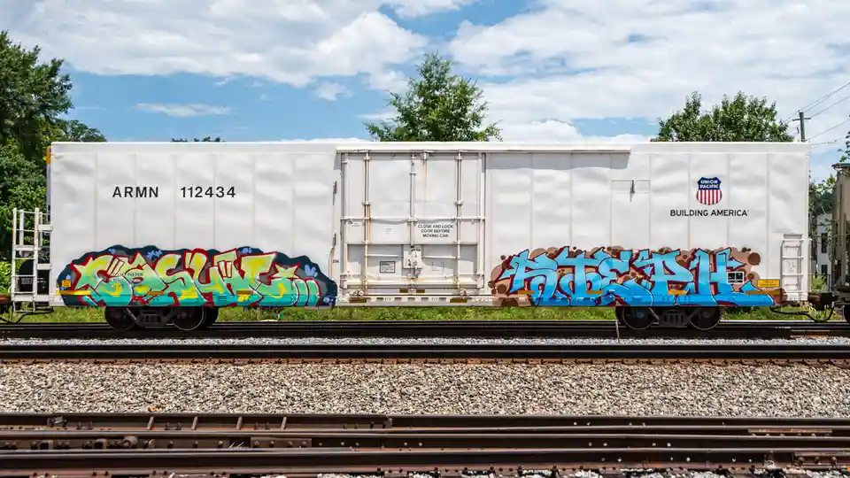 A white Union Pacific freight train car sits on tracks under a partly cloudy sky, featuring large, colorful graffiti tags on…