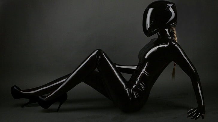 A person in a shiny black latex catsuit and high heels is sitting on a gray floor, wearing a full-face black motorcycle…