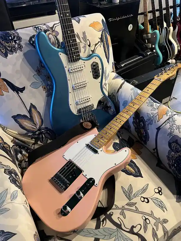 Two electric guitars, one pink and one blue, rest on a floral-patterned couch. The pink guitar is in the foreground with a…