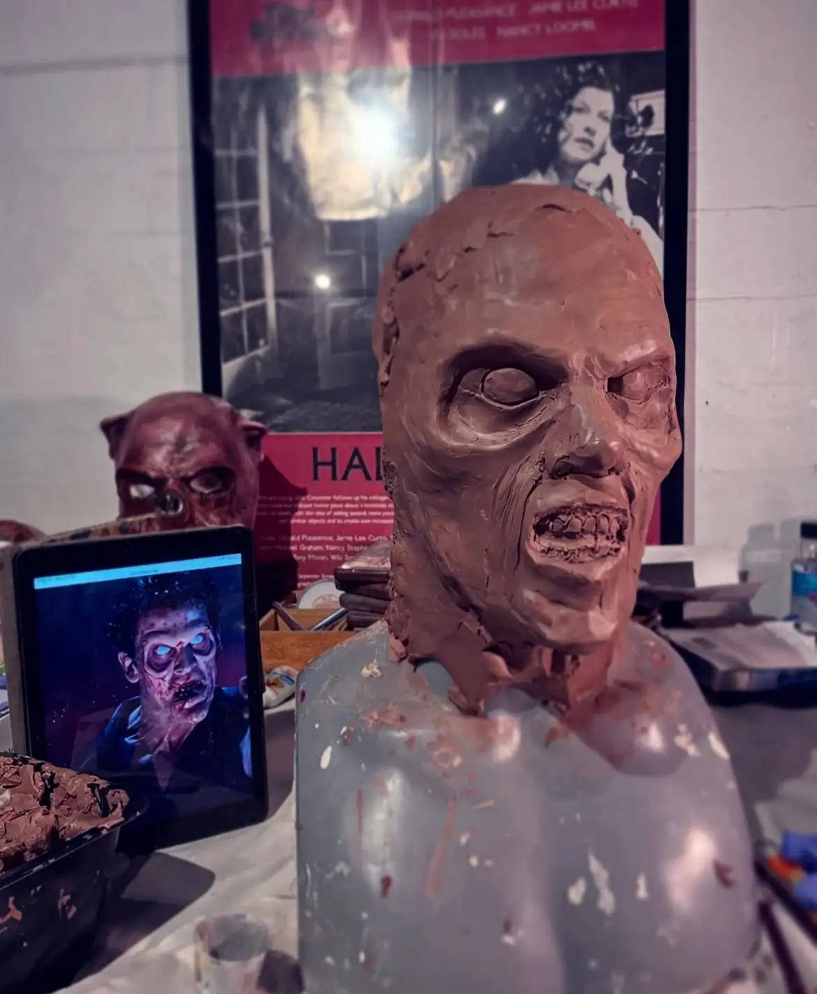A clay sculpture of a zombie head with an open mouth and hollow eyes sits on a workbench, next to a tablet displaying a…