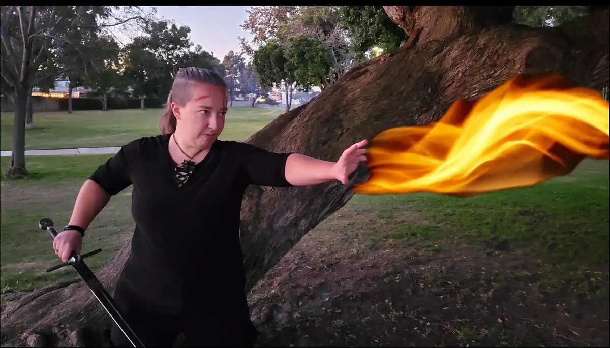 A person in a black shirt stands outdoors next to a large tree, holding a sword in one hand and extending the other toward a…