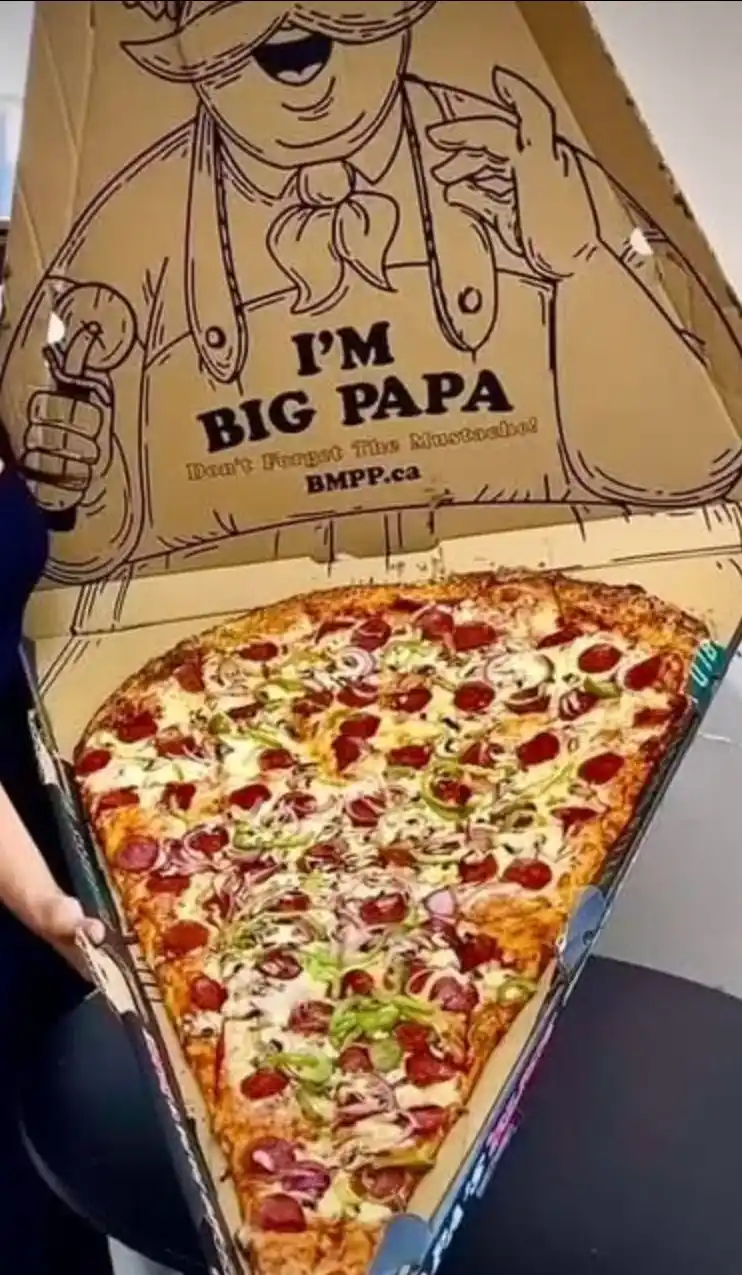 A giant, triangular pizza sits inside an oversized cardboard pizza box featuring a cartoon chef character with a mustache…