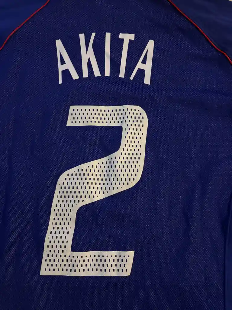 A close-up of the back of a royal blue sports jersey with red trim on the shoulders, featuring the name 'AKITA' in white…