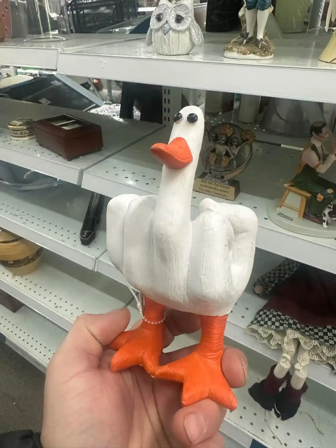 A person holds a white plastic goose figurine with orange beak and feet, but the body is shaped like a hand with fingers…