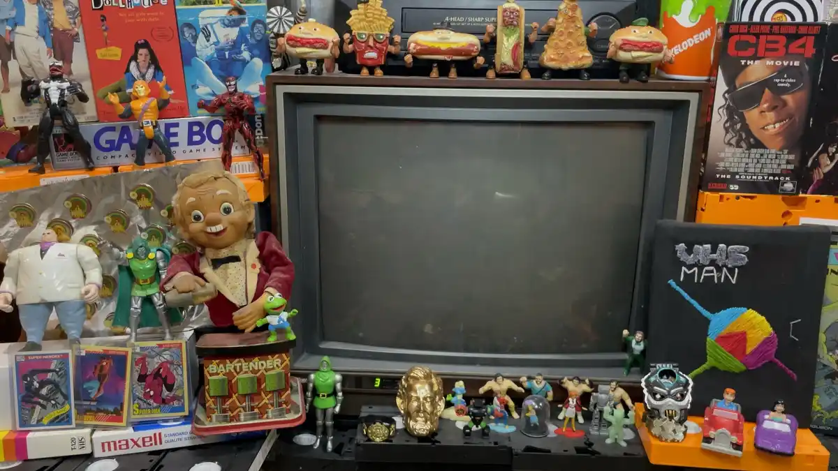 An old CRT television sits in a cluttered display surrounded by vintage toys, action figures, VHS tapes, and movie posters…