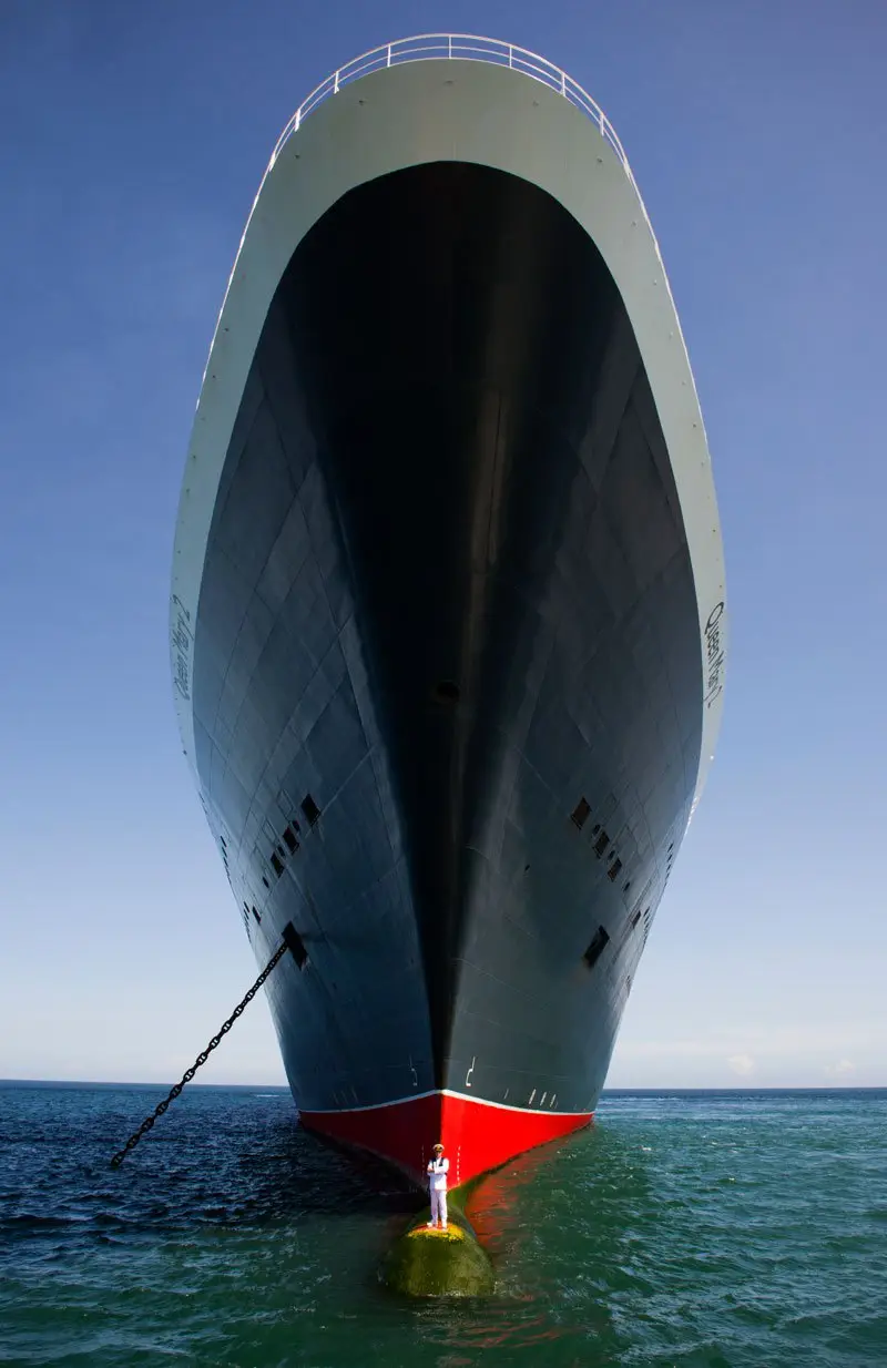 A person in white uniform stands on a small green buoy at the bow of a massive ocean liner, looking up at the ship's…