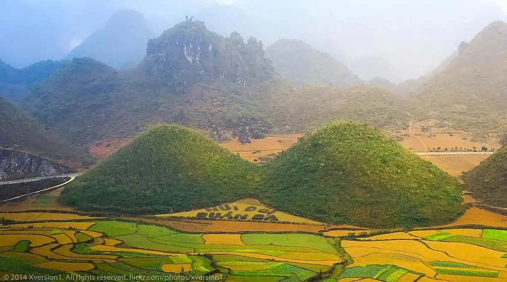 A scenic landscape photo of rolling green hills and terraced rice paddies under a hazy sky, with two prominent rounded hills…