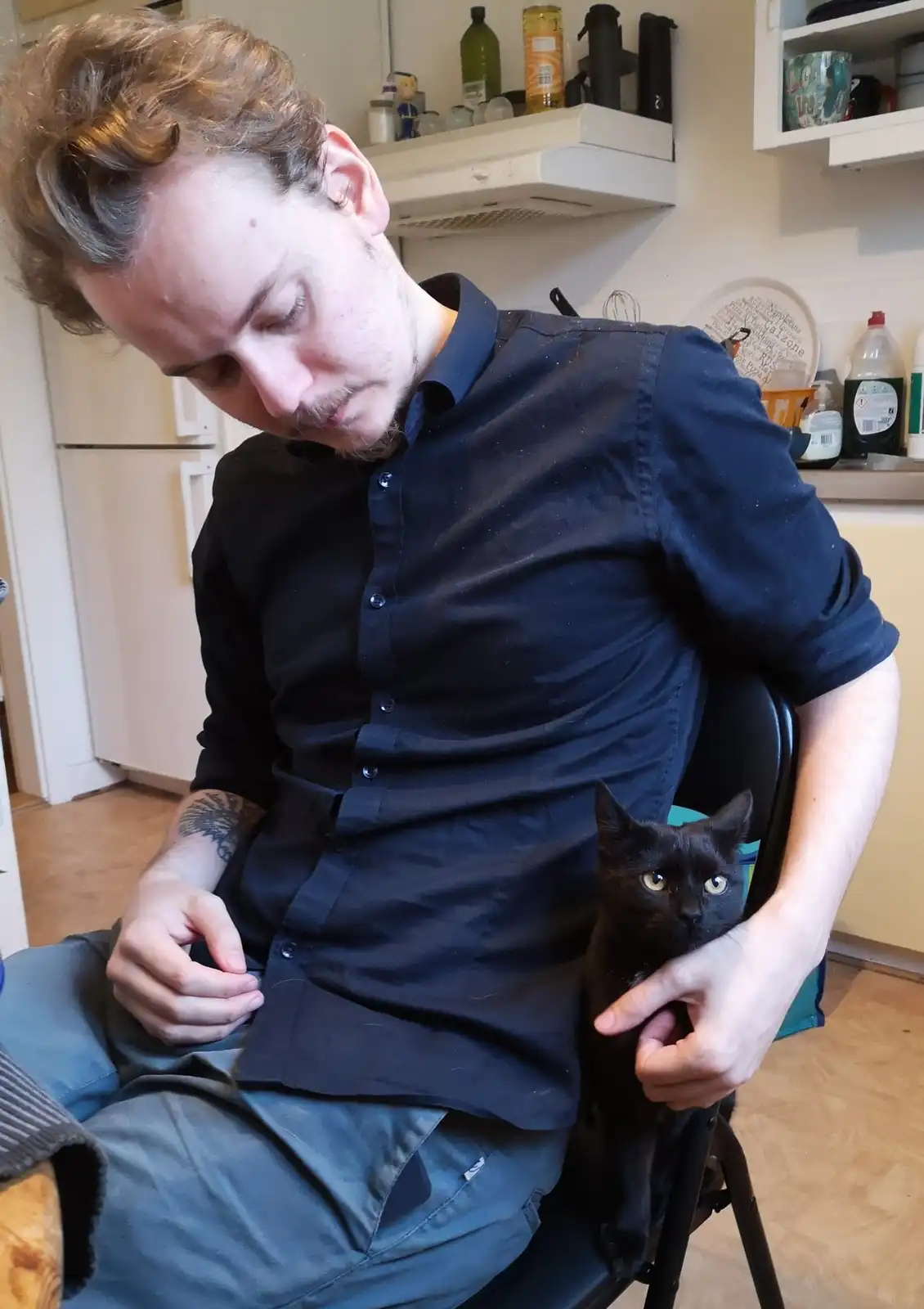 A man with curly hair and a dark shirt sits in a kitchen chair, looking down with a tired expression while holding a black…