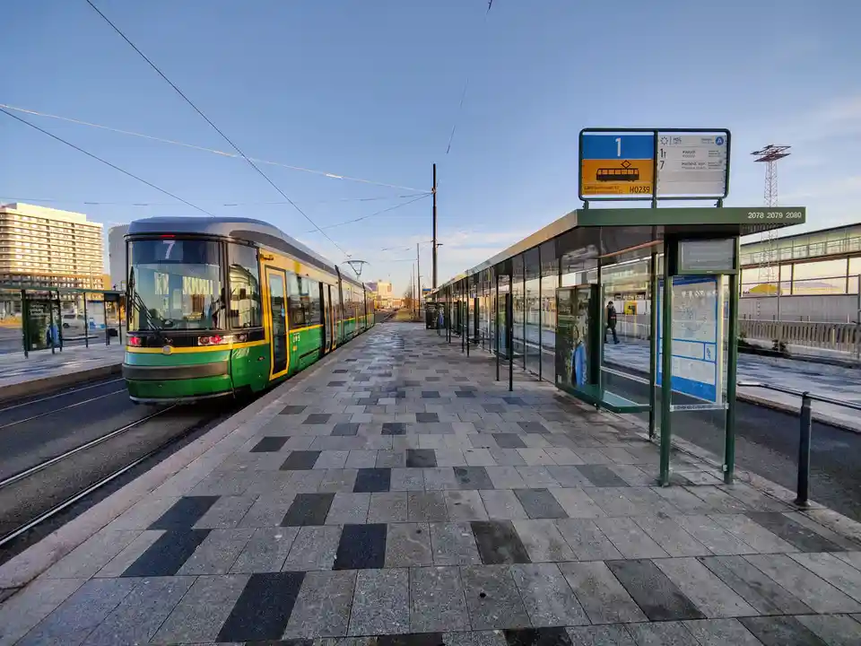 A green and yellow tram, displaying route number 7, is stopped at a modern tram station platform under a clear blue sky. The…