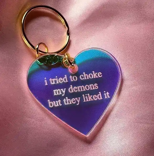 A heart-shaped, iridescent acrylic keychain with a gold ring and small charms rests on pink satin fabric. The keychain has…
