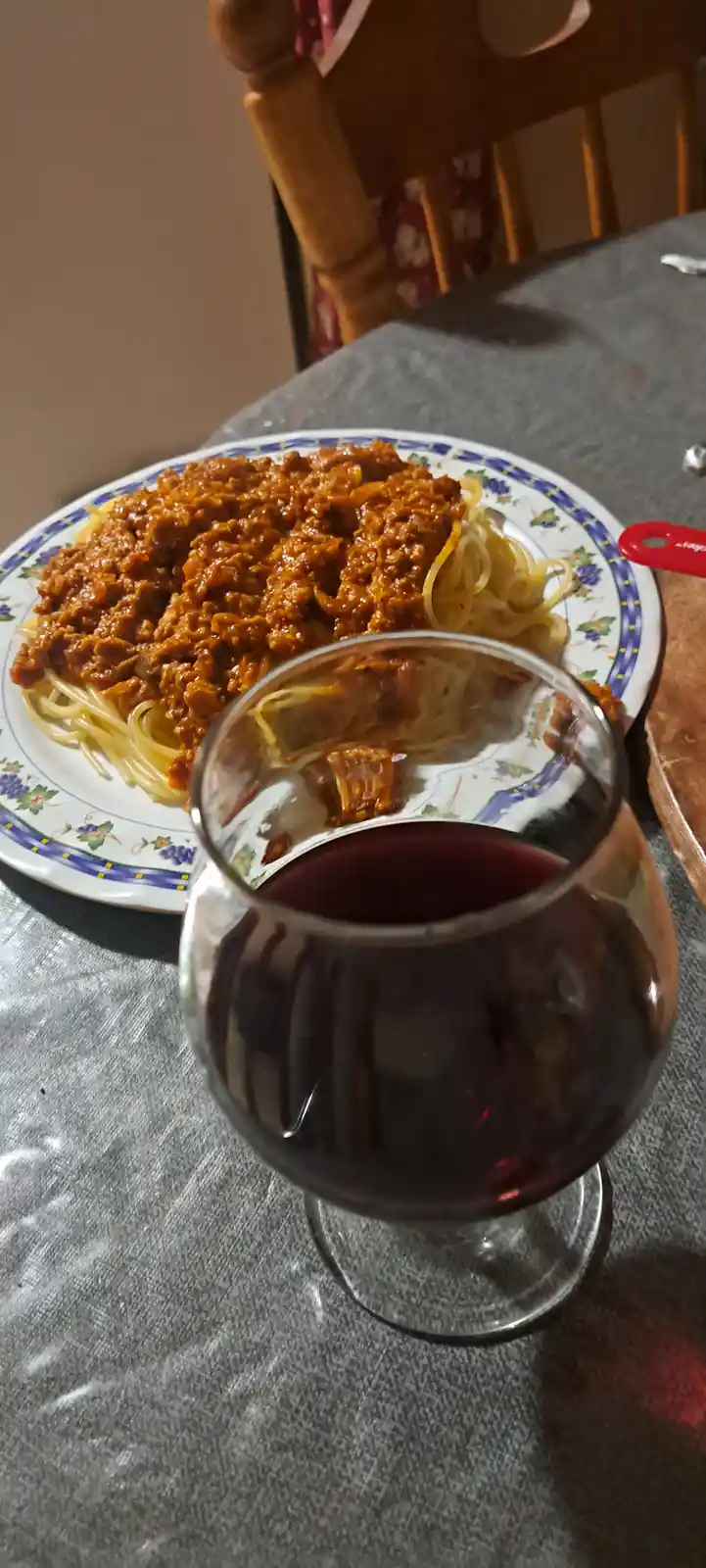 A close-up photo shows a plate of spaghetti covered in a thick meat sauce, with a glass of red wine in the foreground. The…