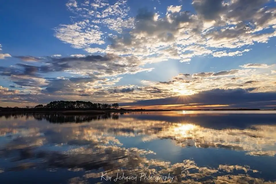 A serene landscape photograph shows a sunrise or sunset over a calm body of water, with the sky filled with scattered clouds…