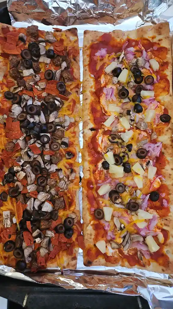 Two rectangular flatbreads on aluminum foil, side-by-side, one topped with meat, mushrooms, and olives, the other with…