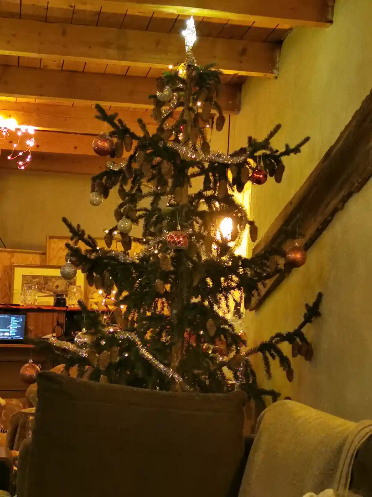 A decorated christmas tree with lights, ornaments, and tinsel stands in a cozy room with wooden beams and furniture. The…