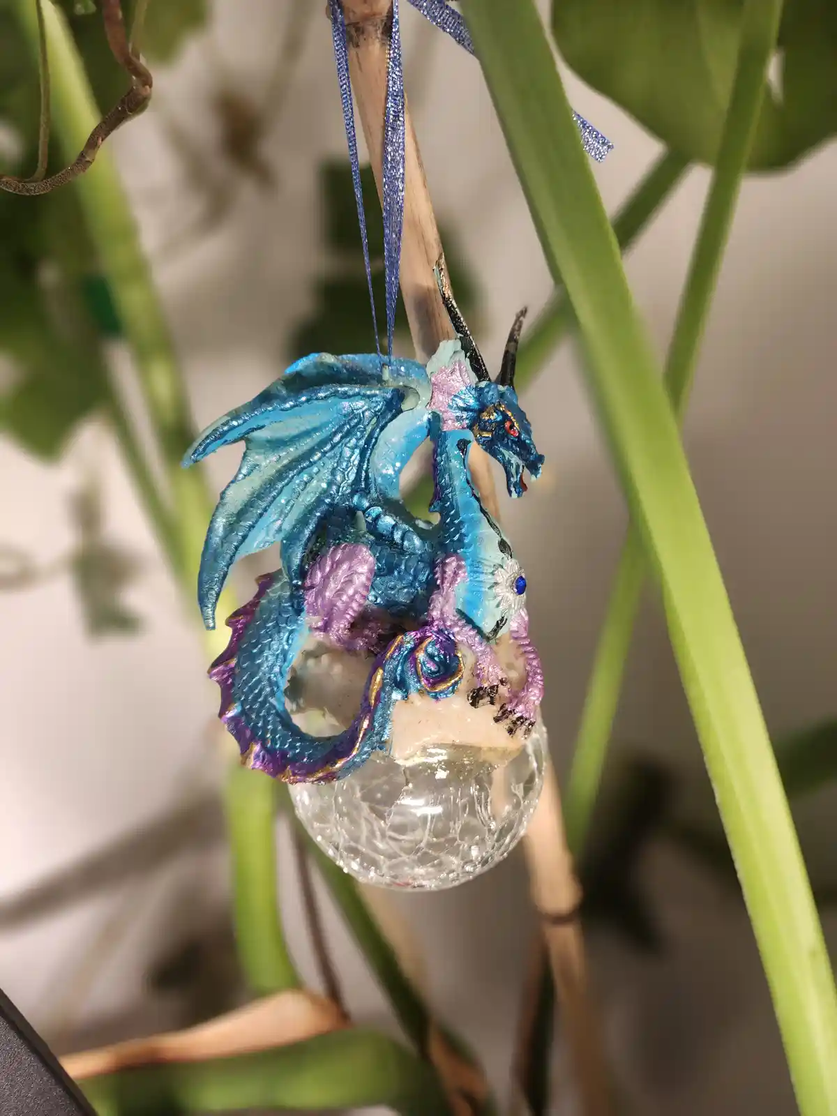 A small, glittery blue dragon ornament with purple accents is hanging from a blue ribbon tied to a wooden stick, positioned…