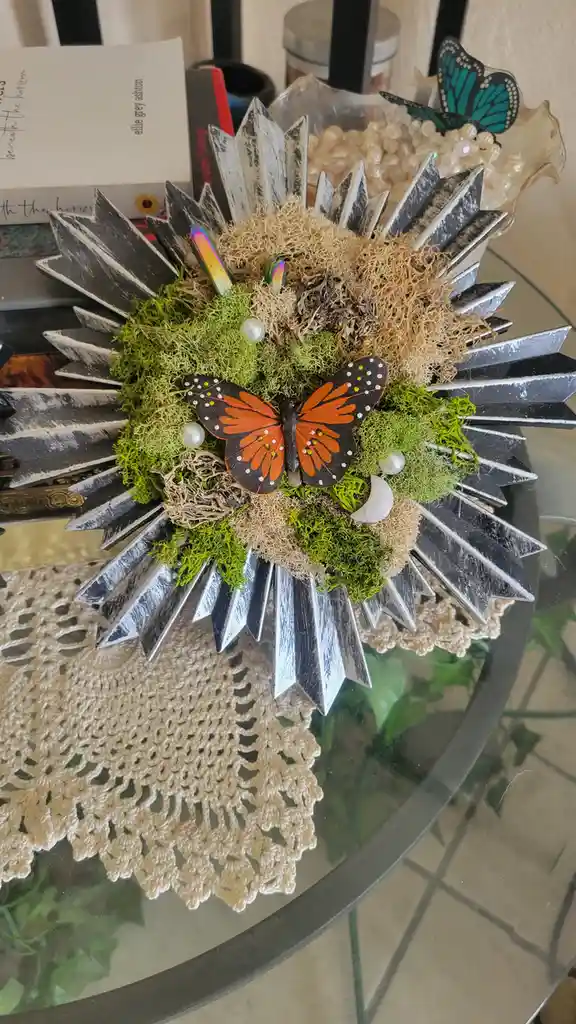 A decorative sunflower-style centerpiece with black-and-white painted wooden rays, green and beige moss, a large orange and…