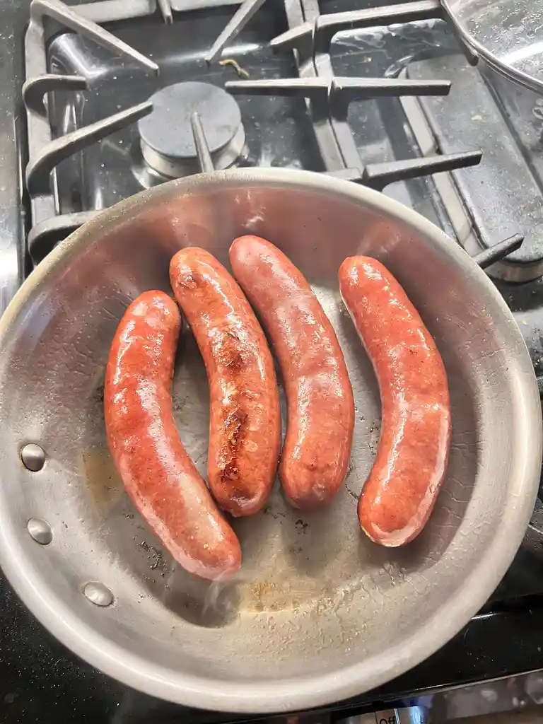 A close-up photo shows four raw-looking sausages in a metal frying pan on a gas stove. The sausages are reddish-pink…