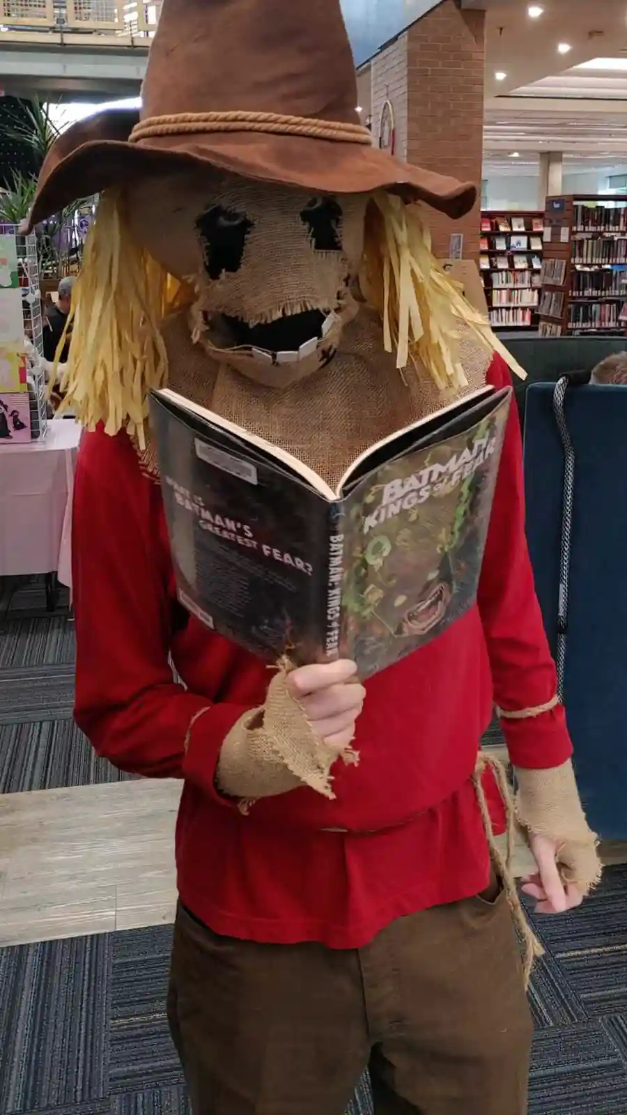 A person dressed as a scarecrow with a burlap mask, straw hair, and a brown hat is standing in a library, holding an open…