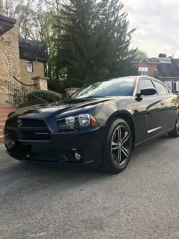 A black Dodge Charger is parked on a residential street in front of a stone house with a staircase. The car has a glossy…