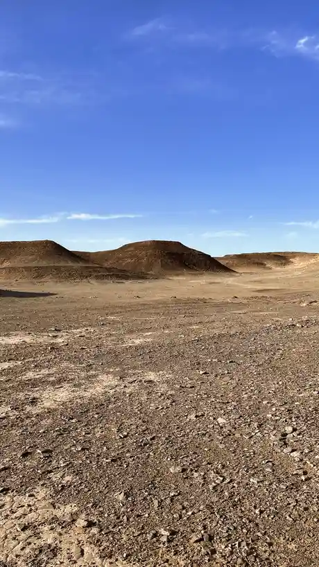 A wide-angle photo of a barren, rocky desert landscape under a clear blue sky with distant low hills. Overlaid text reads…