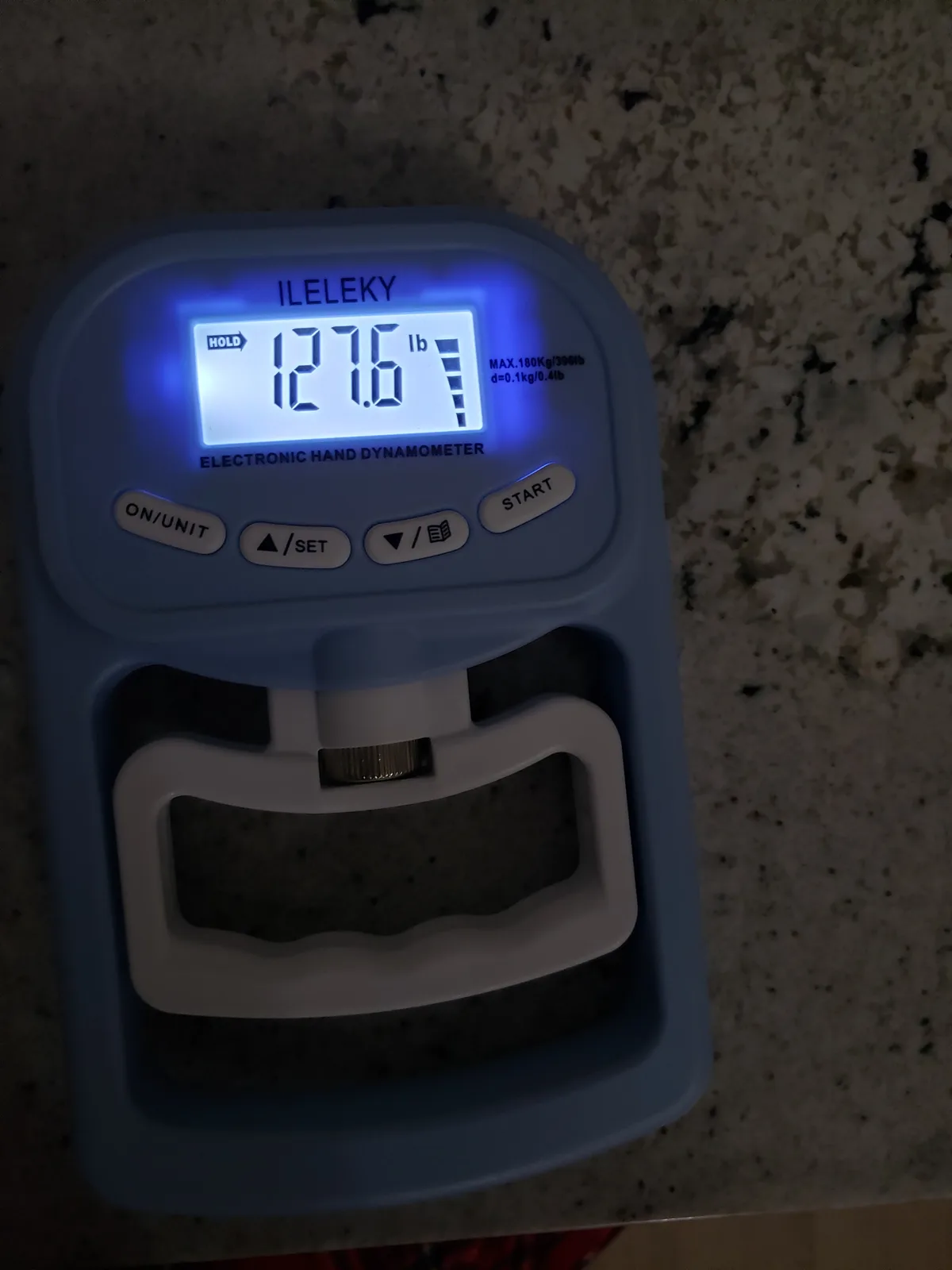A blue ILELEKY electronic hand dynamometer displays '127.6 lb' on its glowing screen, sitting on a speckled countertop. The…