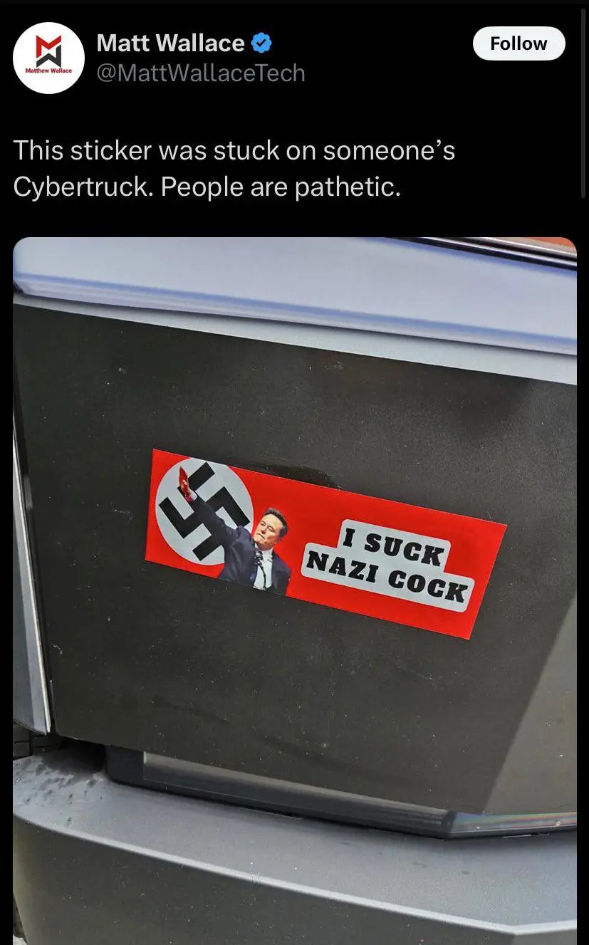 A red rectangular sticker is affixed to the black rear panel of a Tesla Cybertruck. The sticker features a swastika symbol…