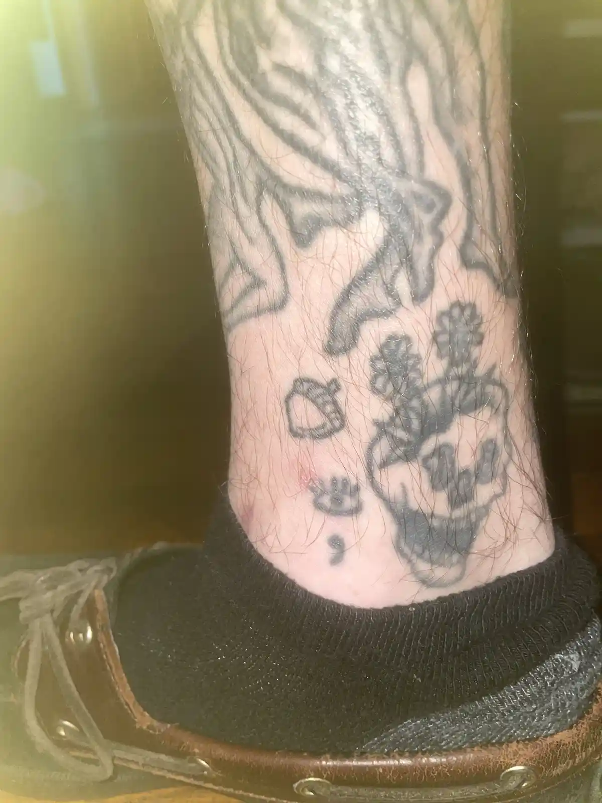 A close-up photo shows the ankle and lower calf of a person wearing a black sock and brown shoe, with several tattoos…
