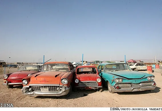 A row of four vintage American cars, mostly red and teal, are parked in a dusty, open-air junkyard under a clear blue sky…