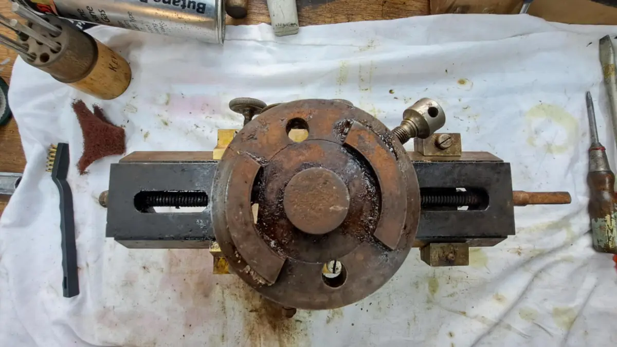 A rusty, worn-out mechanical device, possibly a lathe chuck or similar tool, sits on a stained white cloth surrounded by…