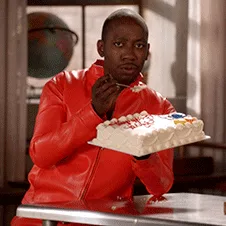 A man in a red jacket sits at a table with a rectangular white cake decorated with red and blue candies. He begins eating…