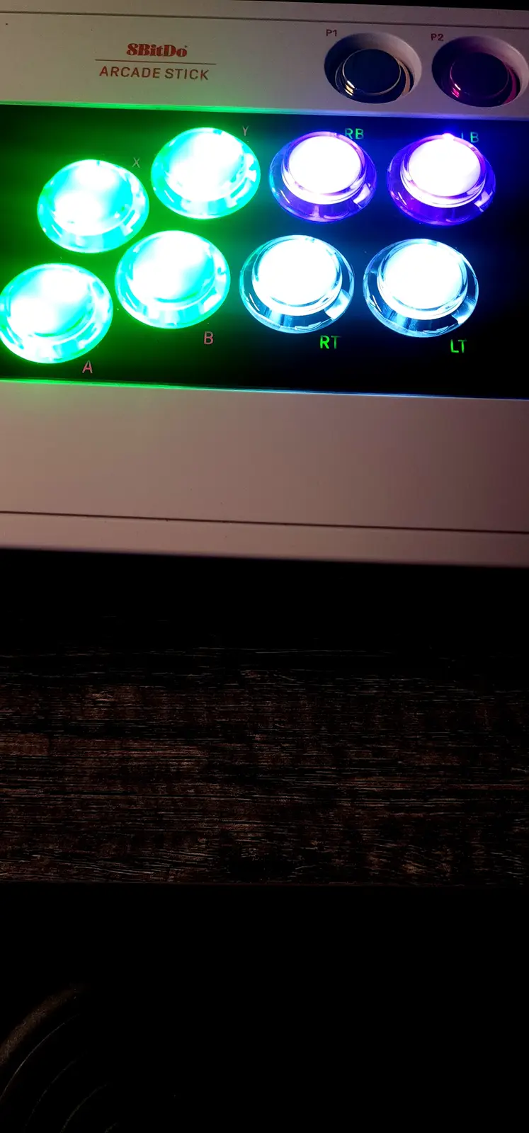 A close-up of an 8BitDo arcade stick with all eight action buttons illuminated in bright green and purple lights. The…