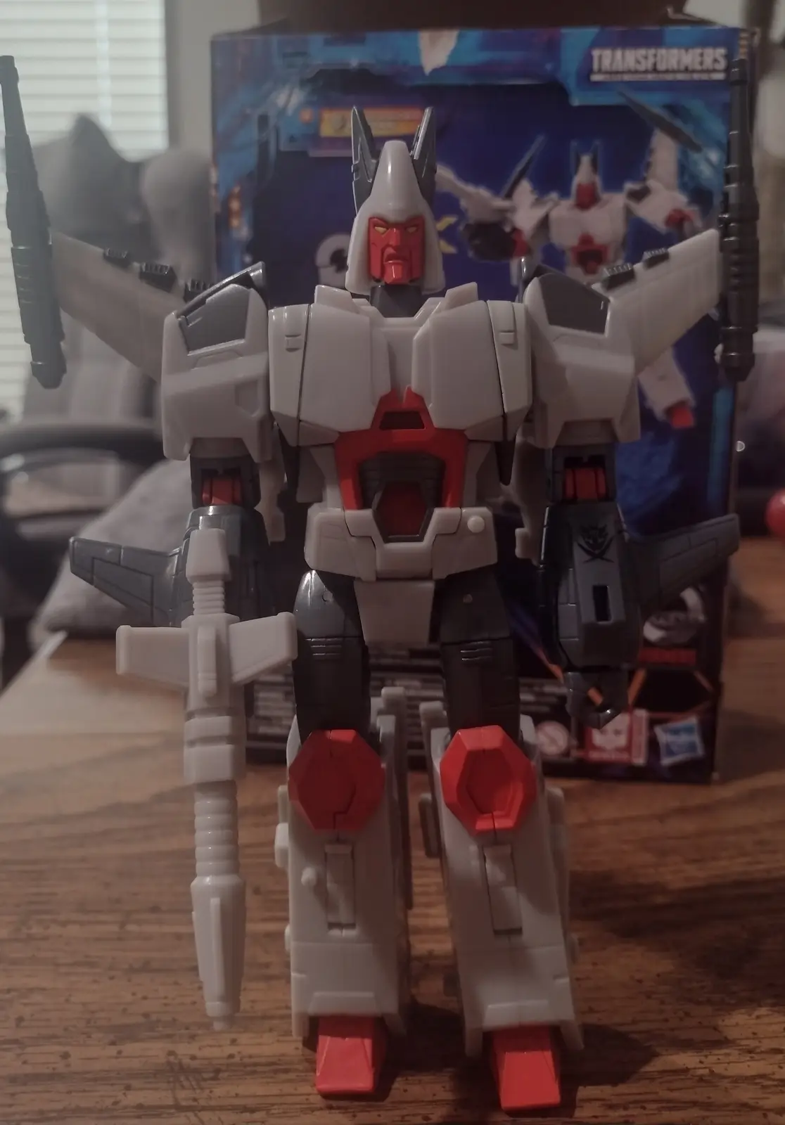 A white, red, and black Transformers action figure stands on a wooden table with its arms outstretched, holding weapons, in…