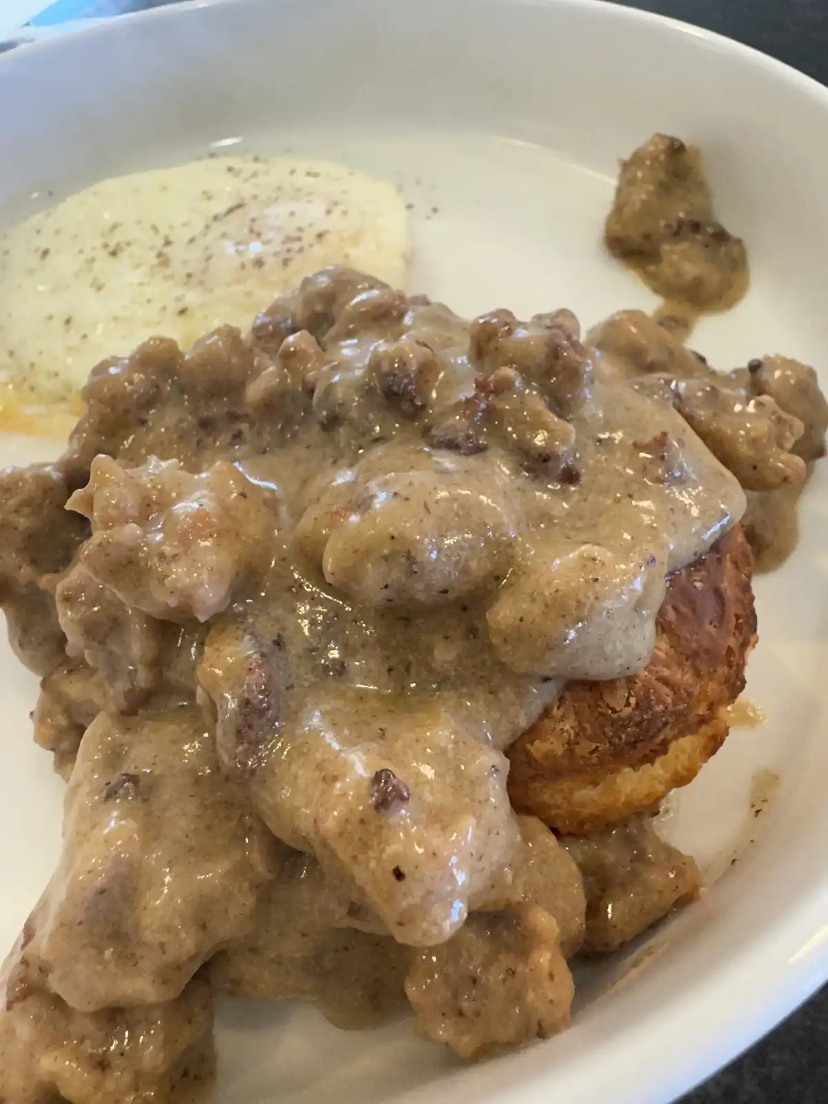 A close-up photo shows a white plate with a golden-brown biscuit smothered in thick, creamy gravy with visible meat chunks…