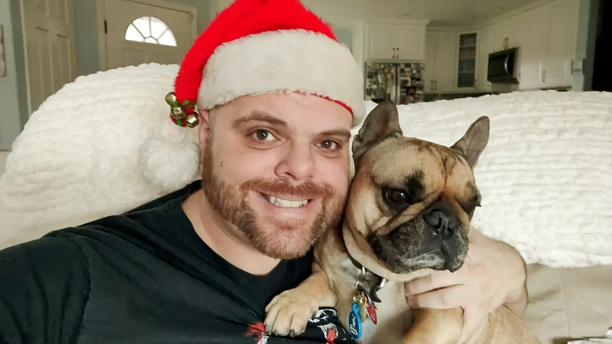 A man with a beard wearing a red and white Santa hat smiles at the camera while holding a French bulldog on his lap. The…