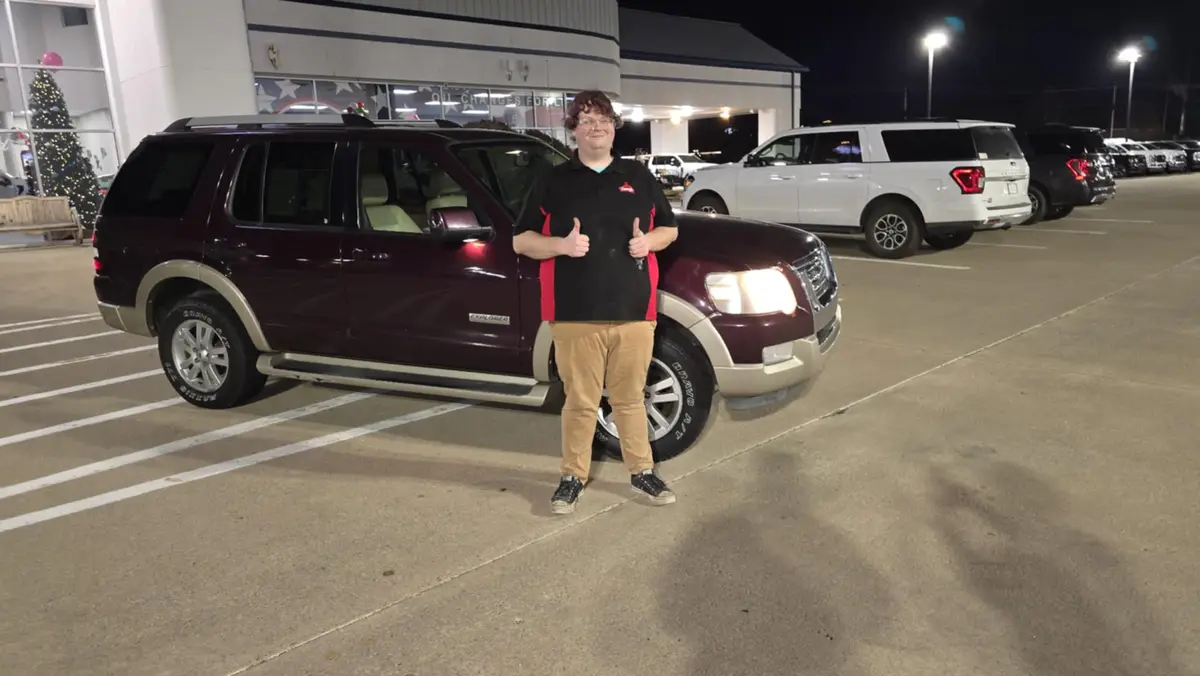 A person stands in a parking lot at night in front of a maroon Ford Explorer, giving two thumbs up. The vehicle is parked in…