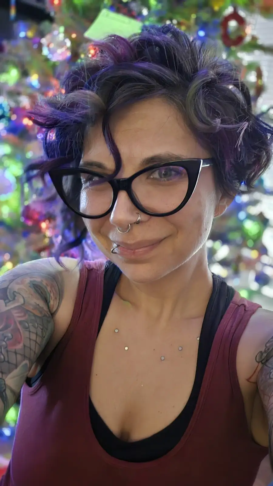 A person with short purple hair, black cat-eye glasses, a nose ring, and multiple chest piercings smiles at the camera while…