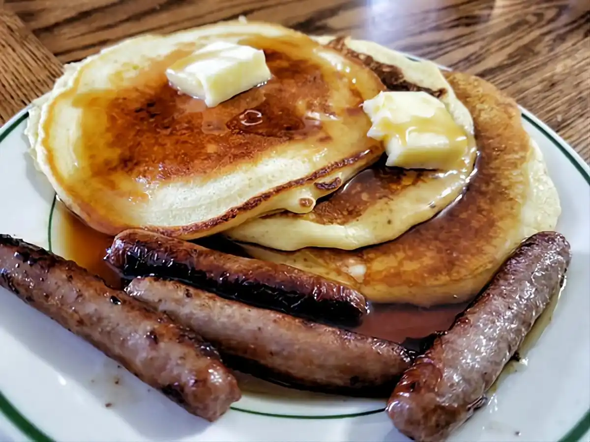 A close-up photo shows a plate of golden-brown pancakes topped with syrup and butter, accompanied by several cooked…