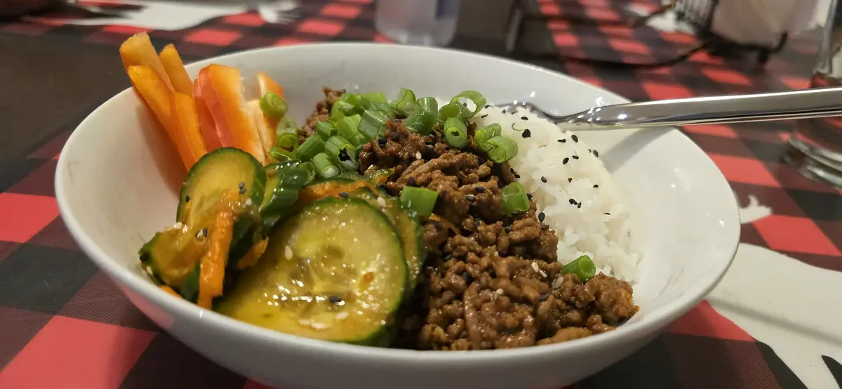A white bowl filled with white rice, brown ground meat, sliced cucumbers, orange bell pepper strips, and chopped green…