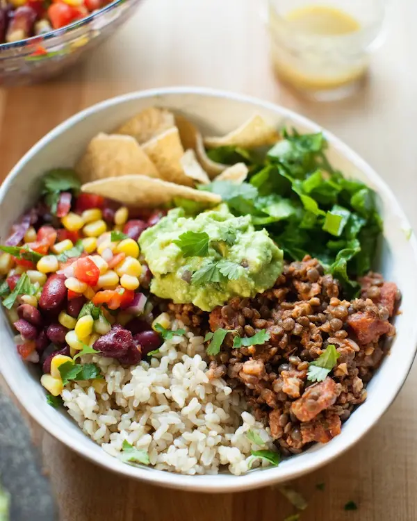 A close-up photo of a colorful, healthy-looking bowl filled with brown rice, black beans, corn, guacamole, chopped cilantro…