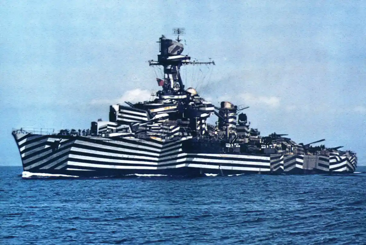 A heavily damaged battleship with black and white stripes painted across its hull, floating in the ocean under a blue sky…
