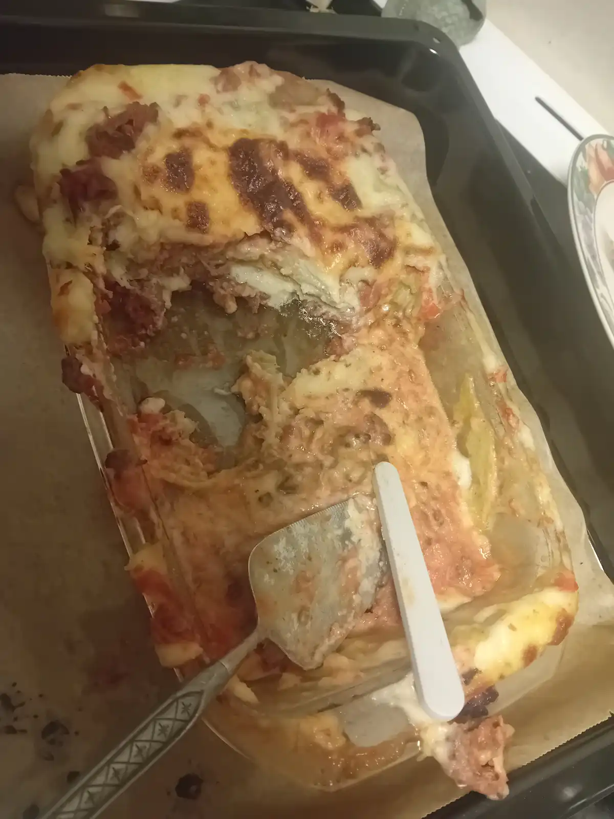 A partially eaten lasagna in a glass baking dish sits on a black tray, with a metal spatula and a white plastic utensil…
