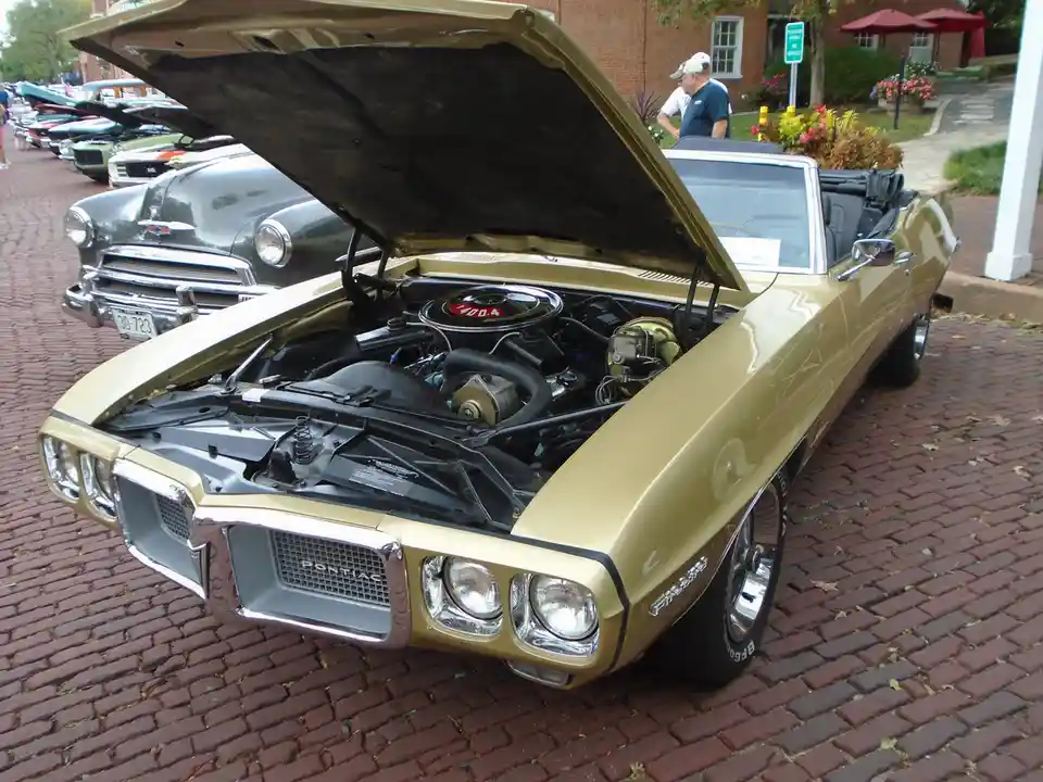 A gold Pontiac GTO convertible with its hood open, revealing a 400 cubic inch engine, parked on a brick street at a car…