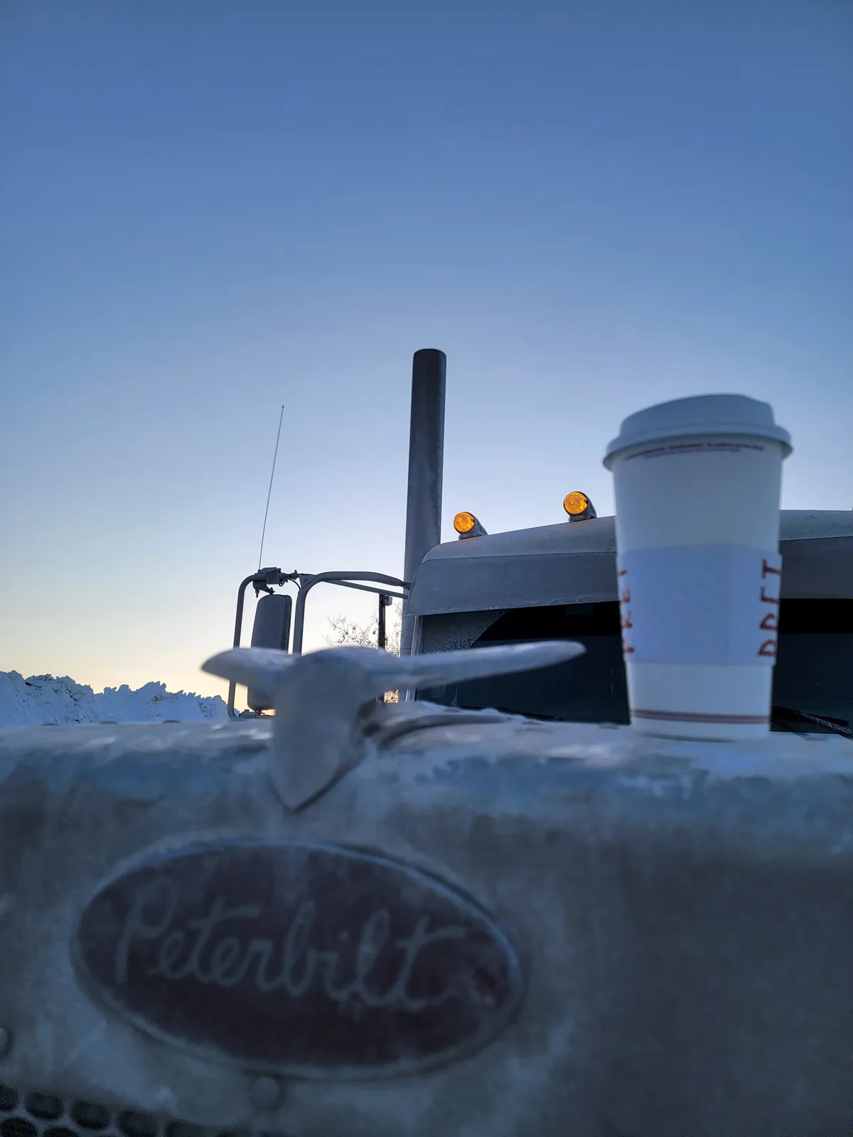 A white disposable coffee cup sits on the icy hood of a Peterbilt semi-truck, with the truck's cab, exhaust stack, and…