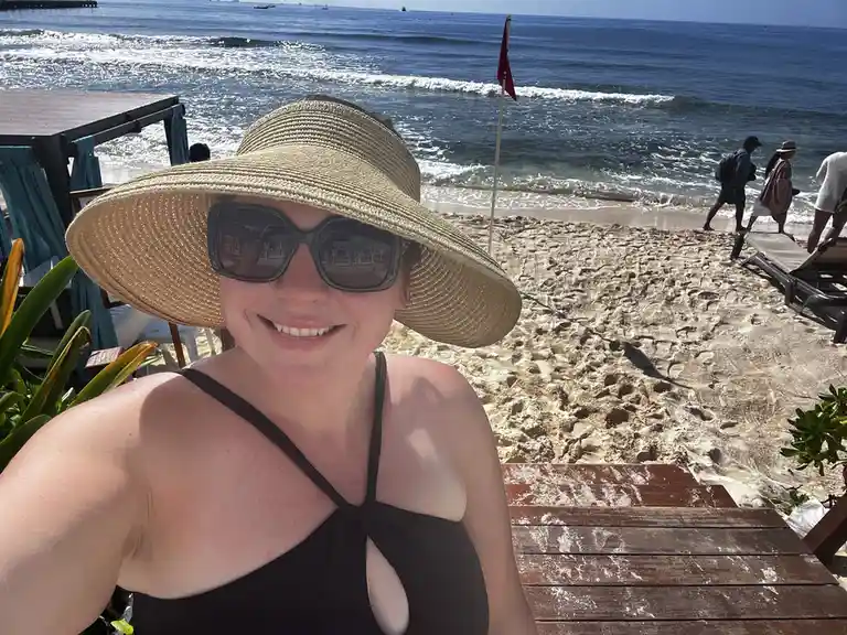 A woman wearing a wide-brimmed straw hat and large sunglasses smiles at the camera while taking a selfie on a wooden…